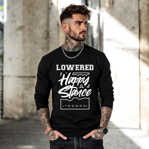 Puede incluir: Sudadera negra de manga larga con el texto blanco "LOWERED Happy Stance". La sudadera tambi&eacute;n tiene el texto "TRDMRK" en un recuadro. La persona lleva una cadena plateada y un reloj.
