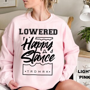 Puede incluir: Sudadera rosa claro con las palabras "LOWERED" encima de "Happy Stance" en una fuente negra estilizada. El texto "TRDMRK" est&aacute; debajo del gr&aacute;fico. La sudadera es de estilo cuello redondo.