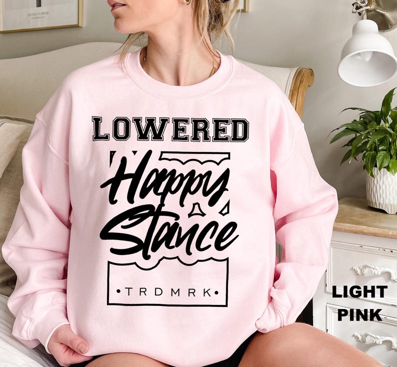 Puede incluir: Sudadera rosa claro con las palabras "LOWERED" encima de "Happy Stance" en una fuente negra estilizada. El texto "TRDMRK" est&aacute; debajo del gr&aacute;fico. La sudadera es de estilo cuello redondo.