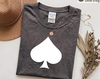 May include: Heather dark grey t-shirt with a white spade graphic. The shirt has a crew neck and short sleeves. A gold-colored necklace with a pendant is draped on the shirt. A straw hat and dried flowers are in the background.