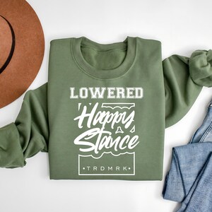Puede incluir: Una sudadera verde oliva con la inscripci&oacute;n blanca "LOWERED Happy Stance". La sudadera est&aacute; doblada, con un sombrero marr&oacute;n y unos vaqueros azules en el fondo. El texto incluye la marca comercial "TRDMRK".