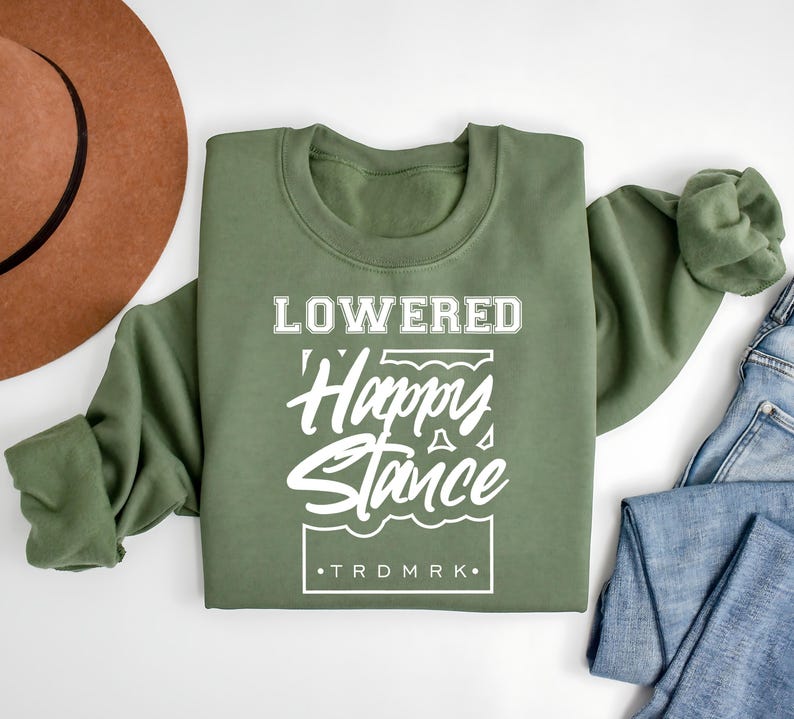 Puede incluir: Una sudadera verde oliva con la inscripci&oacute;n blanca "LOWERED Happy Stance". La sudadera est&aacute; doblada, con un sombrero marr&oacute;n y unos vaqueros azules en el fondo. El texto incluye la marca comercial "TRDMRK".