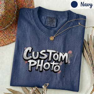 以下が含まれることがあります： ネイビーブルーのクルーネックTシャツ。白の掠れたグラフィティ風フォントで「CUSTOM PHOTO」とプリントされています。襟の内側にはComfort Colorsのタグがあり、生地の上には細いゴールドのネックレスが置かれています。