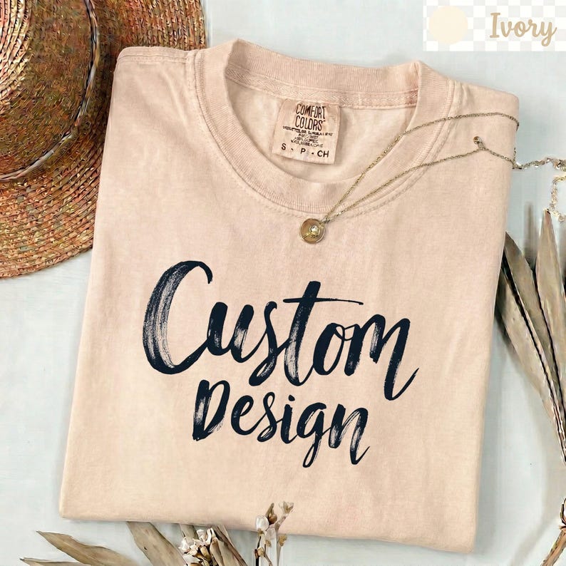 以下が含まれることがあります： アイボリー色の半袖Tシャツで、黒い筆跡スタイルのフォントで「Custom Design」という文字がプリントされています。Tシャツはクルーネックで、襟の内側にComfort Colorsのブランドラベルがあり、細いゴールドトーンのチェーンネックレスが添えられています。