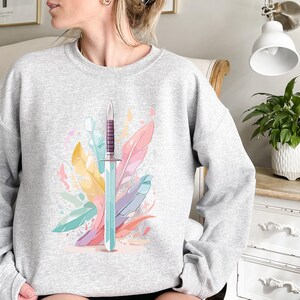 May include: A light gray sweatshirt featuring a watercolor design of a sword and colorful feathers. The sword has a purple handle and a light blue blade. The feathers are in shades of yellow, pink, and blue.