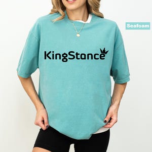 Puede incluir: Camiseta verde mar con el texto negro "KingStance" y un gr&aacute;fico de corona. La camiseta tiene mangas cortas y cuello redondo, y se usa con pantalones cortos negros. Se ve un collar dorado.