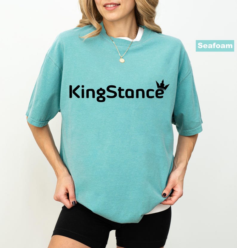 Puede incluir: Camiseta verde mar con el texto negro "KingStance" y un gr&aacute;fico de corona. La camiseta tiene mangas cortas y cuello redondo, y se usa con pantalones cortos negros. Se ve un collar dorado.