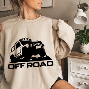 May include: Beige sweatshirt with a black graphic of an off-road vehicle. The vehicle is depicted in silhouette, with the words "OFF ROAD" printed in bold, black letters below the image. The sweatshirt has long sleeves.