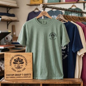 以下が含まれることがあります： セージグリーンの半袖Tシャツ。胸元には木と十字架が描かれた黒い円形のロゴが入っています。手前の木製看板には「OAKWOOD COMMUNITY CHURCH, EST. 2018, SERVE. LOVE. LEAD. CUSTOM GROUP T-SHIRTS BULK ORDERS」という文字が記されています。