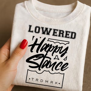 Puede incluir: Camiseta crema doblada con las palabras "LOWERED Happy Stance" en negro y fuente estilizada. El texto est&aacute; dentro de un marco rectangular, con "TRDMRK" en la parte inferior. La camiseta est&aacute; doblada.