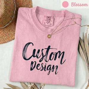 以下が含まれることがあります： クルーネックのライトピンクの半袖Tシャツ。前面には筆致風のフォントで黒い文字でCustom Designとプリントされています。内側の襟元にはComfort Colorsのブランドラベルがあり、サイズSであることが示されています。