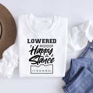 Puede incluir: Una sudadera blanca de manga larga con el texto "LOWERED Happy Stance" en negro. La sudadera se muestra con vaqueros azules y un sombrero marr&oacute;n.