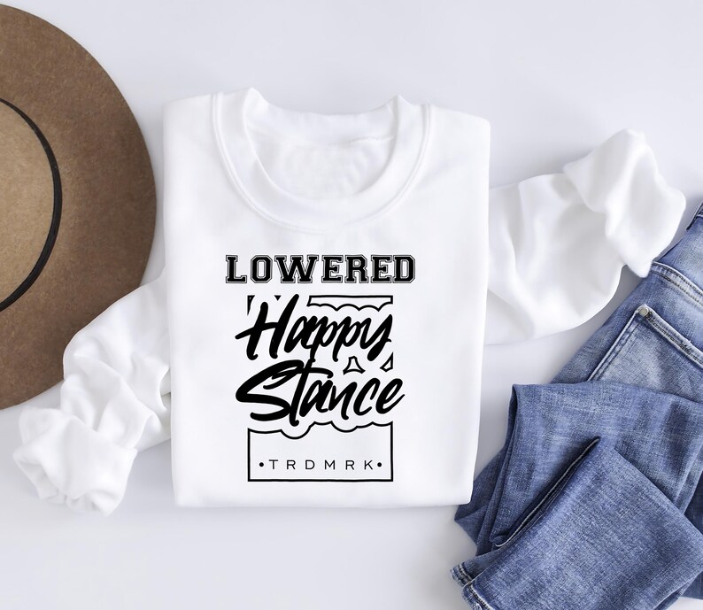 Puede incluir: Una sudadera blanca de manga larga con el texto "LOWERED Happy Stance" en negro. La sudadera se muestra con vaqueros azules y un sombrero marr&oacute;n.