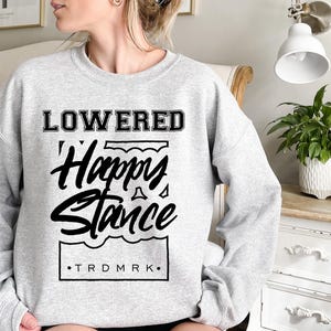 Puede incluir: Sudadera gris claro con la inscripci&oacute;n negra "LOWERED Happy Stance". El texto est&aacute; dentro de un marco rectangular con una l&iacute;nea ondulada en la parte inferior. Las letras "TRDMRK" est&aacute;n en la parte inferior del marco.