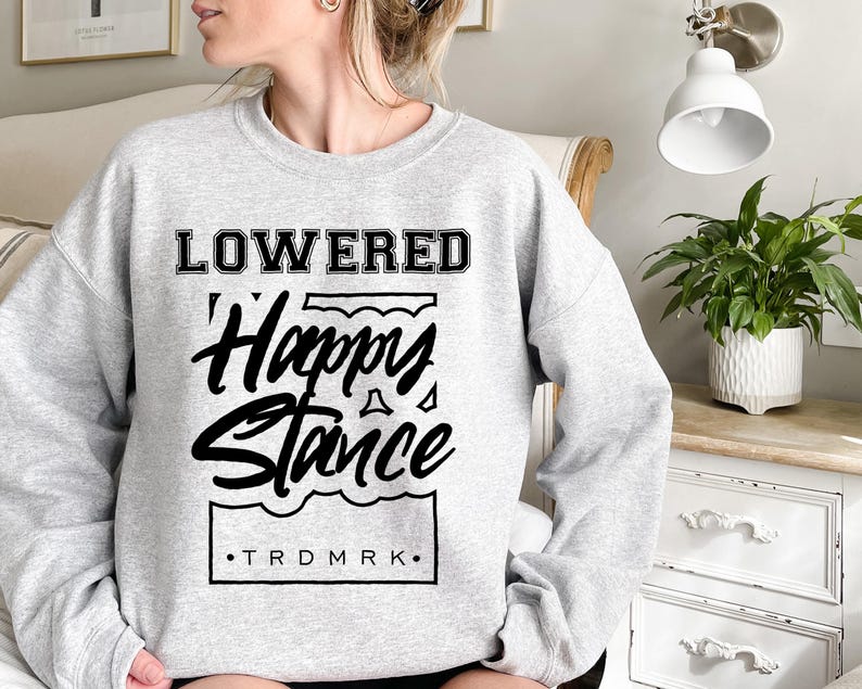 Puede incluir: Sudadera gris claro con la inscripci&oacute;n negra "LOWERED Happy Stance". El texto est&aacute; dentro de un marco rectangular con una l&iacute;nea ondulada en la parte inferior. Las letras "TRDMRK" est&aacute;n en la parte inferior del marco.