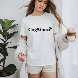 Puede incluir: Una camiseta blanca con el texto negro "KingStance" y un logotipo de corona. La persona lleva una rebeca rosa claro y sostiene un vaso de caf&eacute; en capas con una pajita. Una planta en maceta est&aacute; en el fondo.