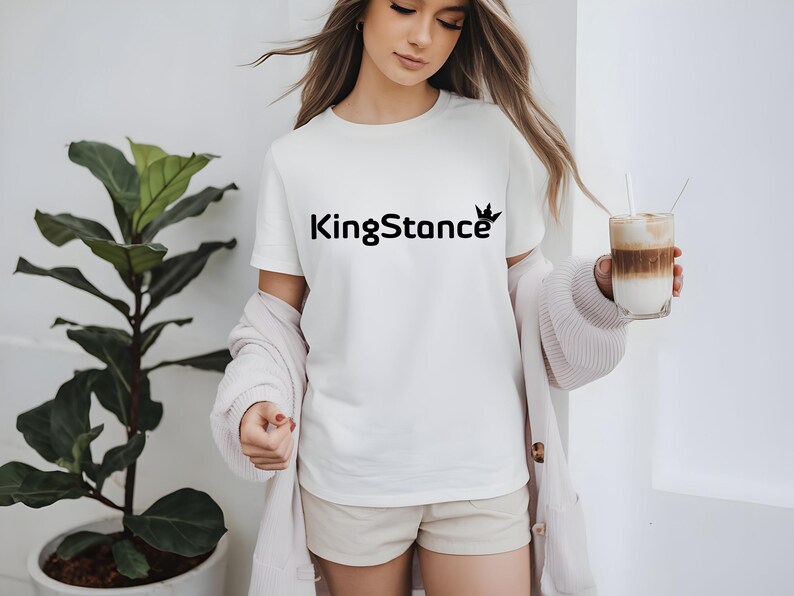 Puede incluir: Una camiseta blanca con el texto negro "KingStance" y un logotipo de corona. La persona lleva una rebeca rosa claro y sostiene un vaso de caf&eacute; en capas con una pajita. Una planta en maceta est&aacute; en el fondo.