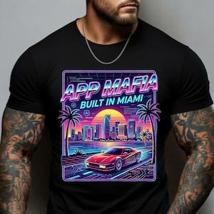 May include: Black t-shirt with a vibrant graphic featuring the text "APP MAFIA BUILT IN MIAMI." The design showcases a red sports car on a road, palm trees, and a city skyline against a sunset backdrop. The design has a retro, neon aesthetic.