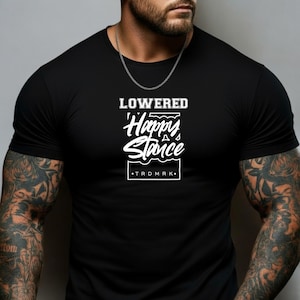 May include: Black t-shirt with the white text "LOWERED Happy Stance" and the brand name "TRDMRK". The shirt has a crew neck and short sleeves. The image shows the shirt on a person.