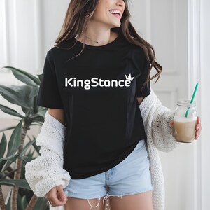 Puede incluir: Camiseta negra con el texto blanco "KingStance" y un gr&aacute;fico de corona. La persona lleva una rebeca blanca y pantalones cortos azules. Sostiene un tarro de cristal con una pajita verde y una bebida beige.
