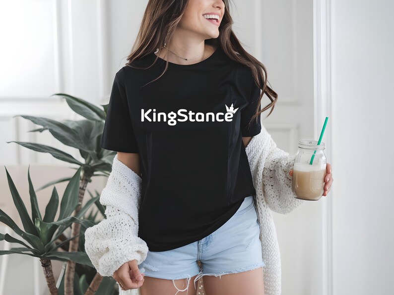 Puede incluir: Camiseta negra con el texto blanco "KingStance" y un gr&aacute;fico de corona. La persona lleva una rebeca blanca y pantalones cortos azules. Sostiene un tarro de cristal con una pajita verde y una bebida beige.