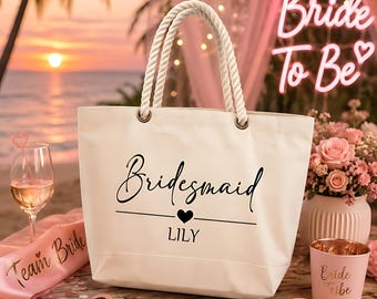 以下が含まれることがあります： 太いロープハンドルが付いた白いキャンバストートバッグ。黒い文字で「Bridesmaid」とプリントされ、その下に小さなハートのアイコンと「LILY」という名前が記されています。バッグはワイングラス、ピンクのサッシュ、ハート型のサングラス、カップと一緒に置かれています。