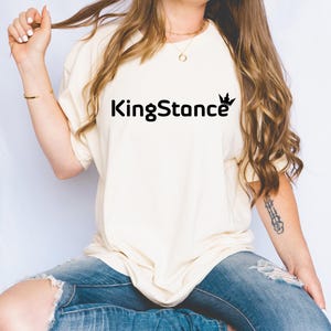 Puede incluir: Camiseta color crema con el texto "KingStance" en negro, con una corona encima de la "ce". La camiseta se combina con vaqueros azules. La modelo lleva un collar y una pulsera dorados.