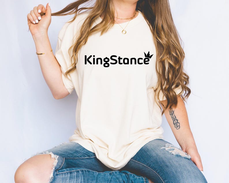 Puede incluir: Camiseta color crema con el texto "KingStance" en negro, con una corona encima de la "ce". La camiseta se combina con vaqueros azules. La modelo lleva un collar y una pulsera dorados.