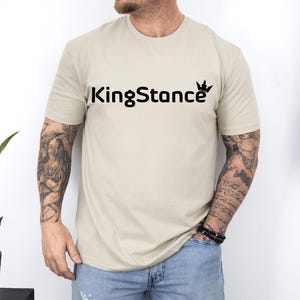 Puede incluir: Una camiseta beige con el texto negro "KingStance" y un gr&aacute;fico de corona. La camiseta de manga corta est&aacute; hecha de un material suave. La persona que lleva la camiseta tiene tatuajes en los brazos.