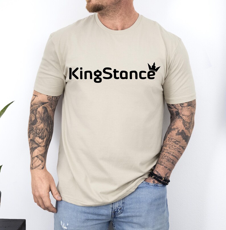 Puede incluir: Una camiseta beige con el texto negro "KingStance" y un gr&aacute;fico de corona. La camiseta de manga corta est&aacute; hecha de un material suave. La persona que lleva la camiseta tiene tatuajes en los brazos.