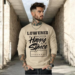 Puede incluir: Una sudadera beige con las palabras "LOWERED Happy Stance" en letras cursivas y en bloque negras. La sudadera tambi&eacute;n tiene el texto "TRDMRK" en la parte inferior. La camisa de manga larga es de color neutro.