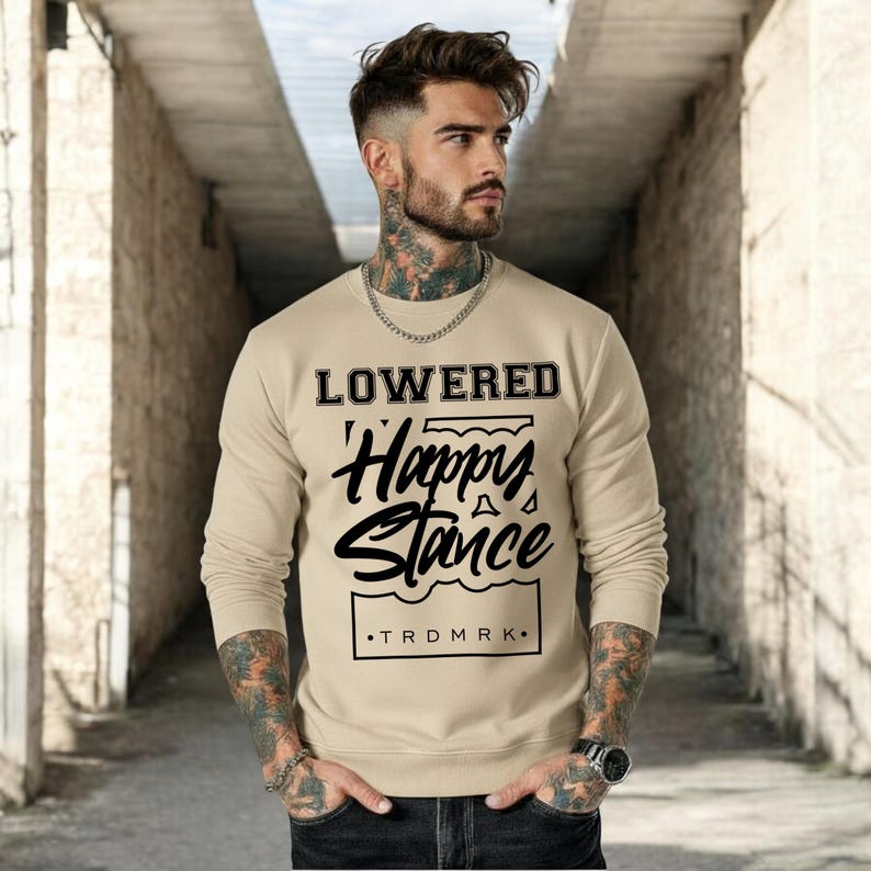 Puede incluir: Una sudadera beige con las palabras "LOWERED Happy Stance" en letras cursivas y en bloque negras. La sudadera tambi&eacute;n tiene el texto "TRDMRK" en la parte inferior. La camisa de manga larga es de color neutro.