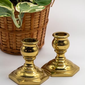 May include: Two gold metal candle holders with a hexagonal base and a round, bulbous center section. The candle holders are on a white surface.