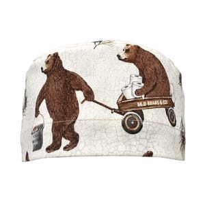 May include: A white scrub cap with a repeating pattern of brown bears. One bear pulls a bucket, and another rides in a wooden wagon filled with toilet paper. The text "M.B. BEARS & CO." is on the wagon.