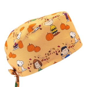 May include: A yellow and orange scrub hat with a pattern of Peanuts characters, pumpkins, and autumn leaves. The hat has a tie on the side.