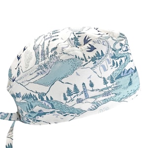 May include: A scrub cap featuring a white background with a detailed blue and teal landscape print. The pattern includes illustrations of a lighthouse, mountains, a bridge, trees, birds, and a sailboat, with tie-back straps at the base.