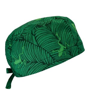 May include: Green scrub cap with a tropical leaf pattern and light green frog illustrations. The cap has a tie closure at the back. The fabric is a vibrant green with dark green leaf veins.