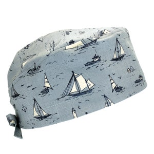 May include: A light blue scrub cap featuring a repeating pattern of dark blue sailboats, lighthouses, and anchors. The cap has a rounded crown, a wide front band, and fabric ties at the back for an adjustable fit, showcasing a nautical theme.