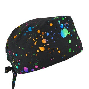 May include: A black surgical scrub cap with a colorful splatter paint design. The paint splatters are in shades of blue, green, orange, yellow, and purple. The cap has a tie in the back for an adjustable fit.