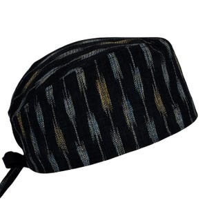 May include: A black scrub cap featuring a vertical ikat-style pattern with alternating streaks of muted gold, light blue, and white. The head covering has a rounded crown, a structured front band, and fabric ties extending from the back for adjustment.