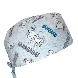 May include: Light blue scrub cap featuring a repeating pattern of Snoopy characters in various poses, with the text "HAHAHAHA!" in blue. The cap has a tie closure at the back and is made of a soft fabric.