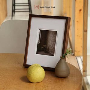 May include: A framed photograph with a dark wood frame and white matting. A small ceramic vase with a sprig of flowers and a yellow fruit sit on a wooden table. The text "LONGMO ART" is in the upper right corner.