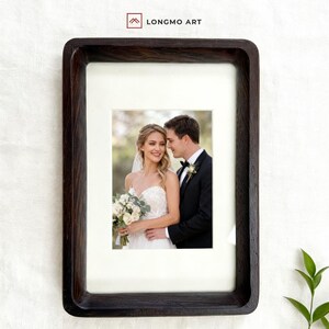 Puede incluir: Un marco de fotos de madera marrón oscuro con una fotografía de una novia y un novio. El marco tiene forma rectangular con esquinas redondeadas. La fotografía muestra a una pareja sonriente con atuendo de boda, la novia sostiene un ramo.