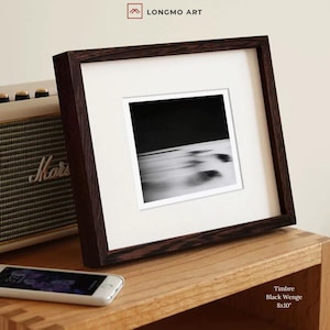 Op de afbeelding: Een ingelijste zwart-wit abstracte kunstafdruk. De donkerbruine lijst omringt een witte passe-partout en een zwart-wit afbeelding. De tekst "Timbre Black Wenge 8x10" staat rechtsonder. De lijst staat op een houten oppervlak.