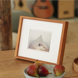 Op de afbeelding: Een vierkante houten fotolijst met een witte passe-partout, met een foto van mensen op een berg. De lijst is gemaakt van lichtbruin hout. De afbeelding is 15 x 20 cm.