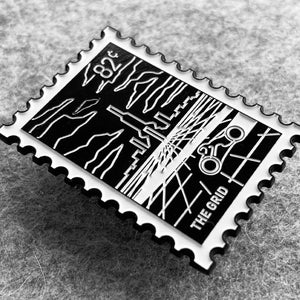 May include: Black and white enamel pin shaped like a postage stamp with a city skyline and a bicycle inside a circle. The text "THE GRID" is at the bottom of the pin.
