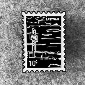 Puede incluir: Pin de esmalte blanco y negro con un diseño vintage de un letrero que dice "East Van St." con un paisaje urbano en el fondo. El pin tiene un borde blanco con un borde festoneado y dice "10¢" en la esquina inferior izquierda.