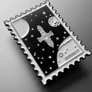 Op de afbeelding: Een zwart-witte emaille pin in de vorm van een postzegel met een ruimteschip in de ruimte met planeten en sterren.
