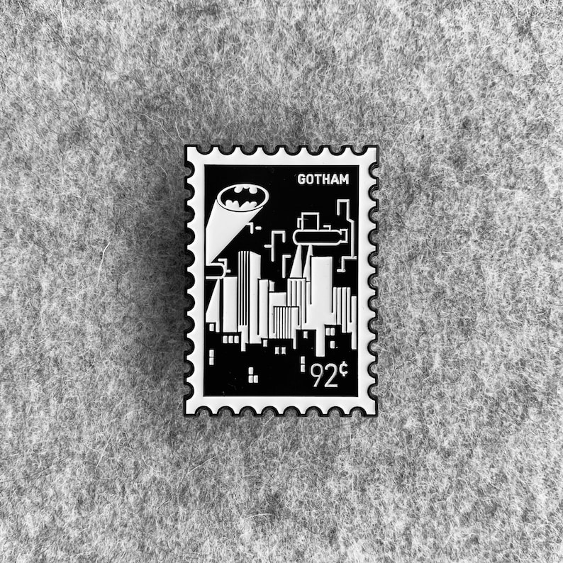 May include: Black and white enamel pin featuring a stylized cityscape with the text "GOTHAM" and "92&cent;" on a black background. The pin is shaped like a postage stamp with a white border.