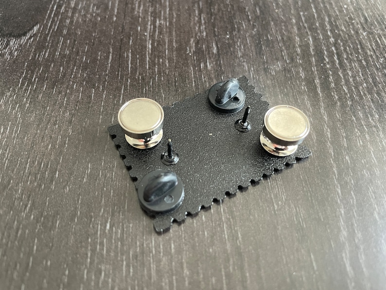 May include: The back of a black enamel pin with a silver metal pin back and a black rubber clutch. The pin is shaped like a rectangle with rounded corners and has a scalloped edge.